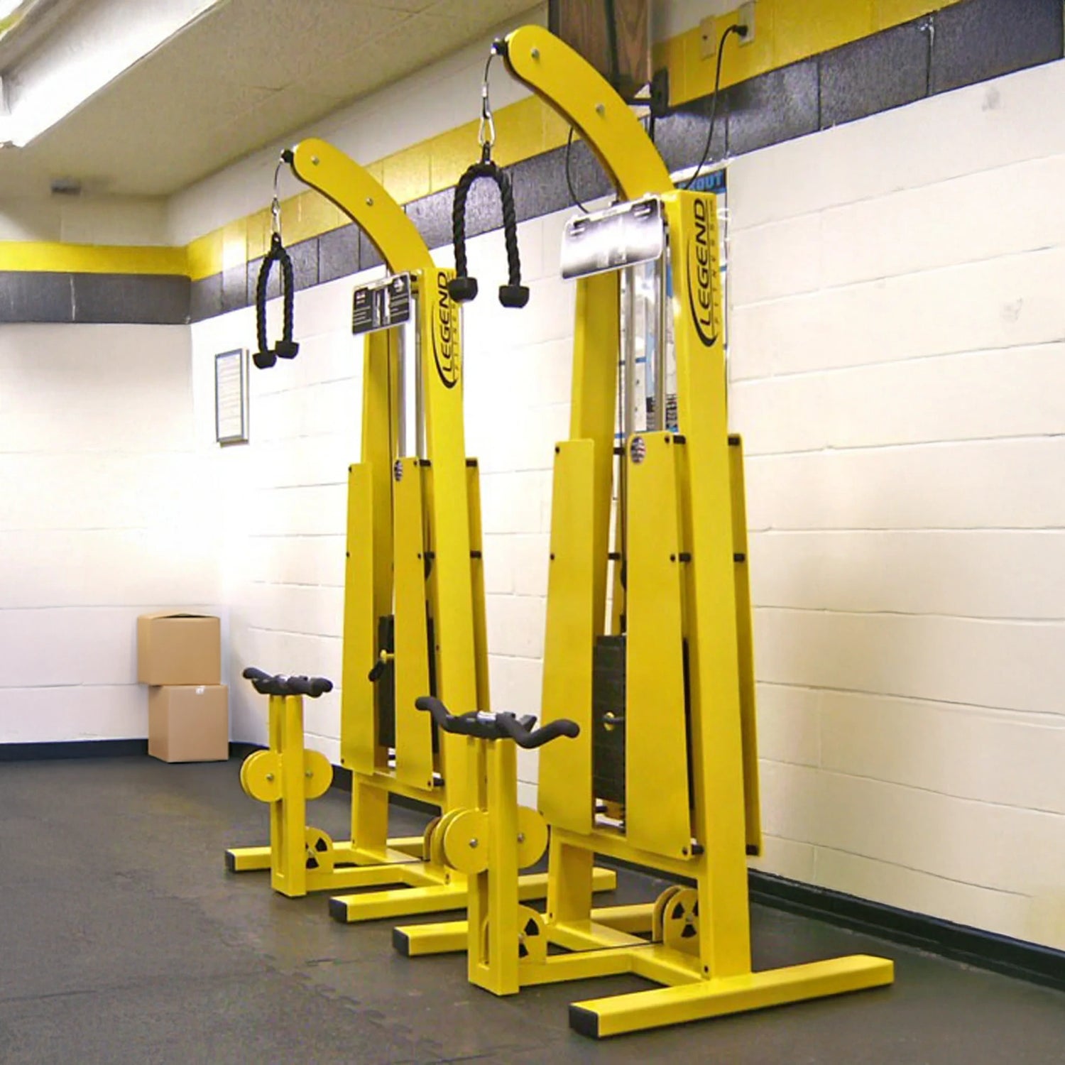 Two Legend Fitness 946 Standing Bicep / Tricep Combo Machines, tall and bright yellow with black handles, stand side by side against a white and yellow gym wall. Each features seats, pulleys, and boxes stacked in the corner.