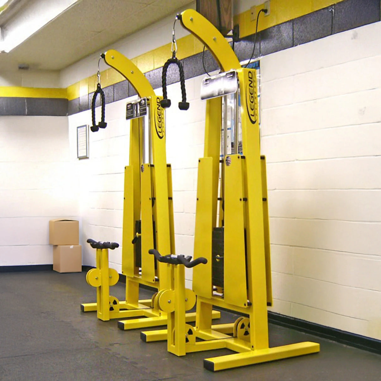 Two Legend Fitness 946 Standing Bicep / Tricep Combo Machines, tall and bright yellow with black handles, stand side by side against a white and yellow gym wall. Each features seats, pulleys, and boxes stacked in the corner.