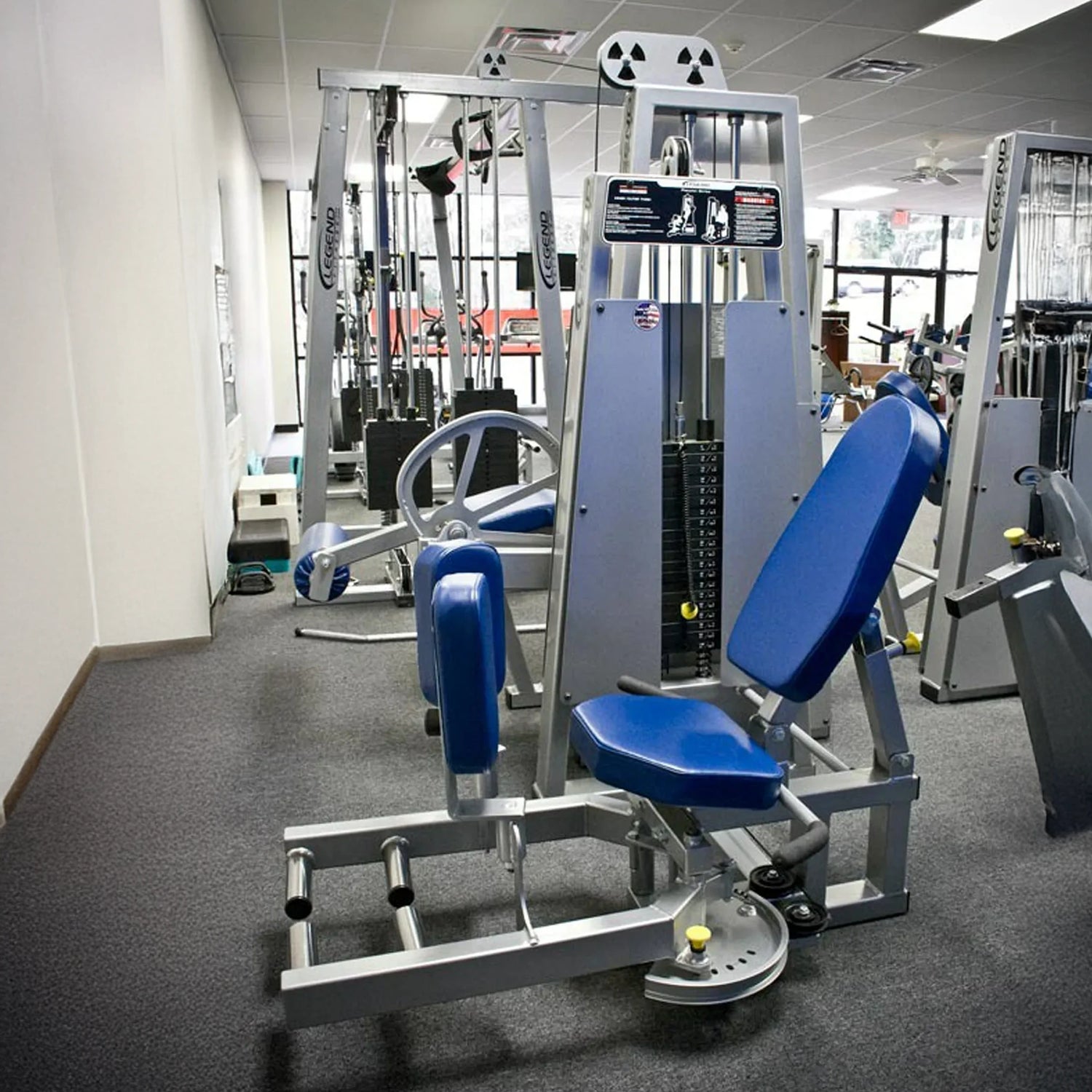 The Legend Fitness 950 Outer Thigh Machine by Legend Fitness, in blue and silver, is positioned in a gym with gray carpet, surrounded by other exercise equipment and large windows that let in natural light.