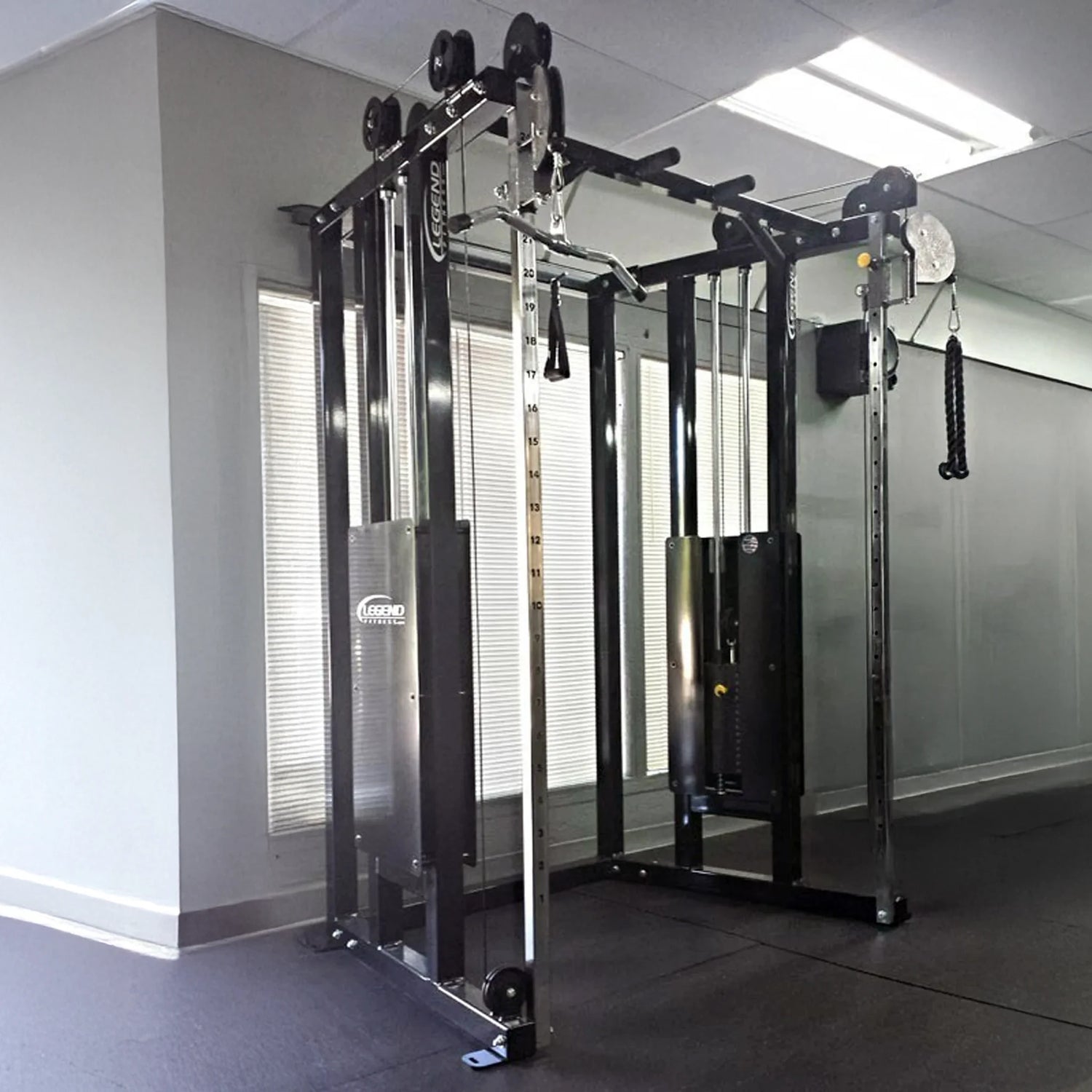 The Legend Fitness 953 Functional Trainer Machine by Legend Fitness stands in a gym corner with large windows and bright lights, featuring adjustable pulleys, weight stacks, and various attachments for strength training.