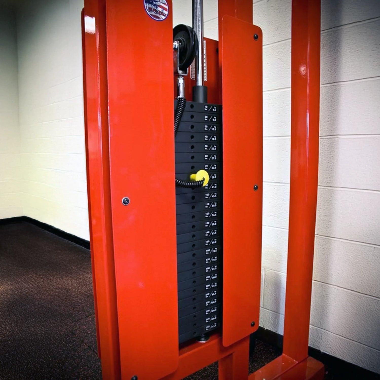 A close-up of the Legend Fitness 953 Functional Trainer Machine by Legend Fitness, featuring black rectangular weight plates with a yellow selector pin, set against a white and beige wall and brown carpeted floor.