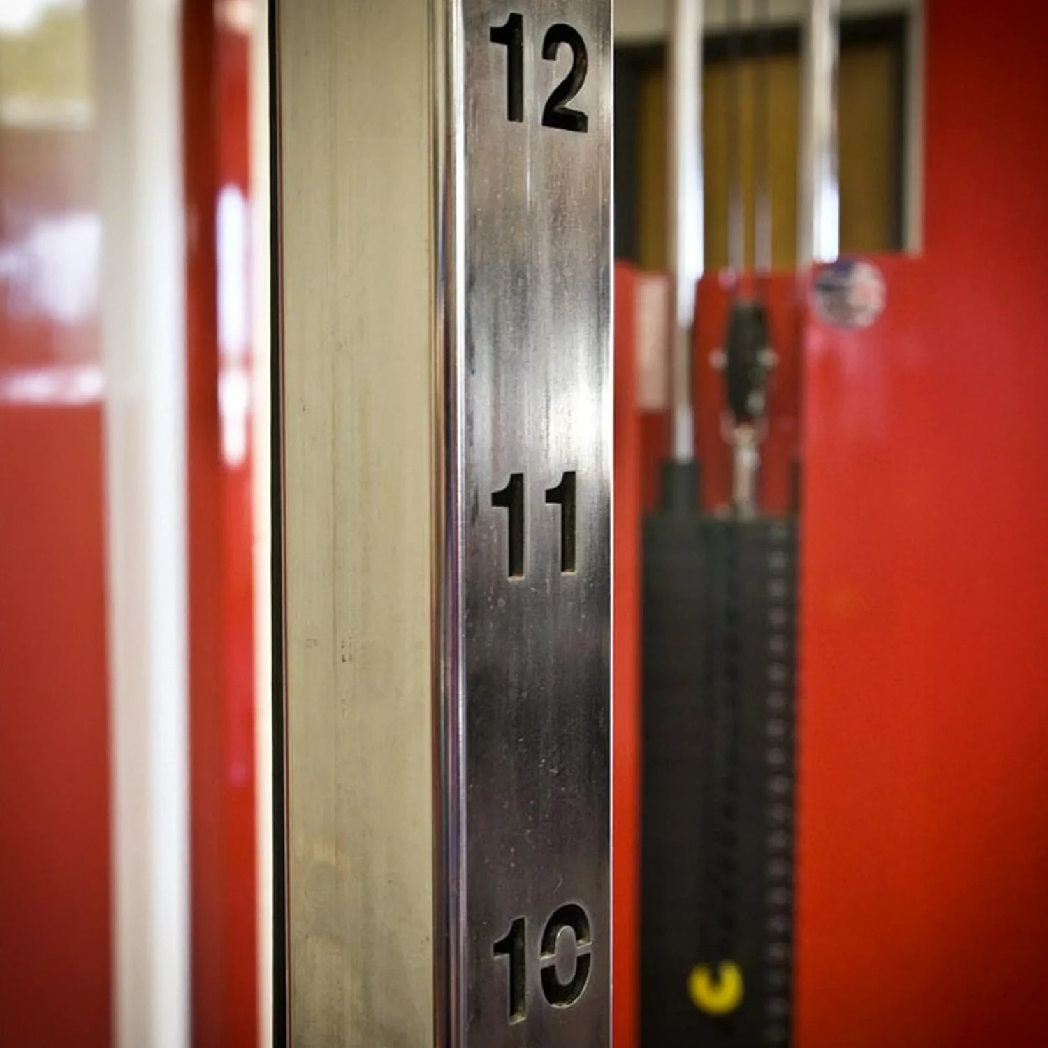 Close-up of a Legend Fitness 953 Functional Trainer Machine by Legend Fitness, showing numbered height markings 10, 11, and 12. The blurred background features red walls and gym equipment.