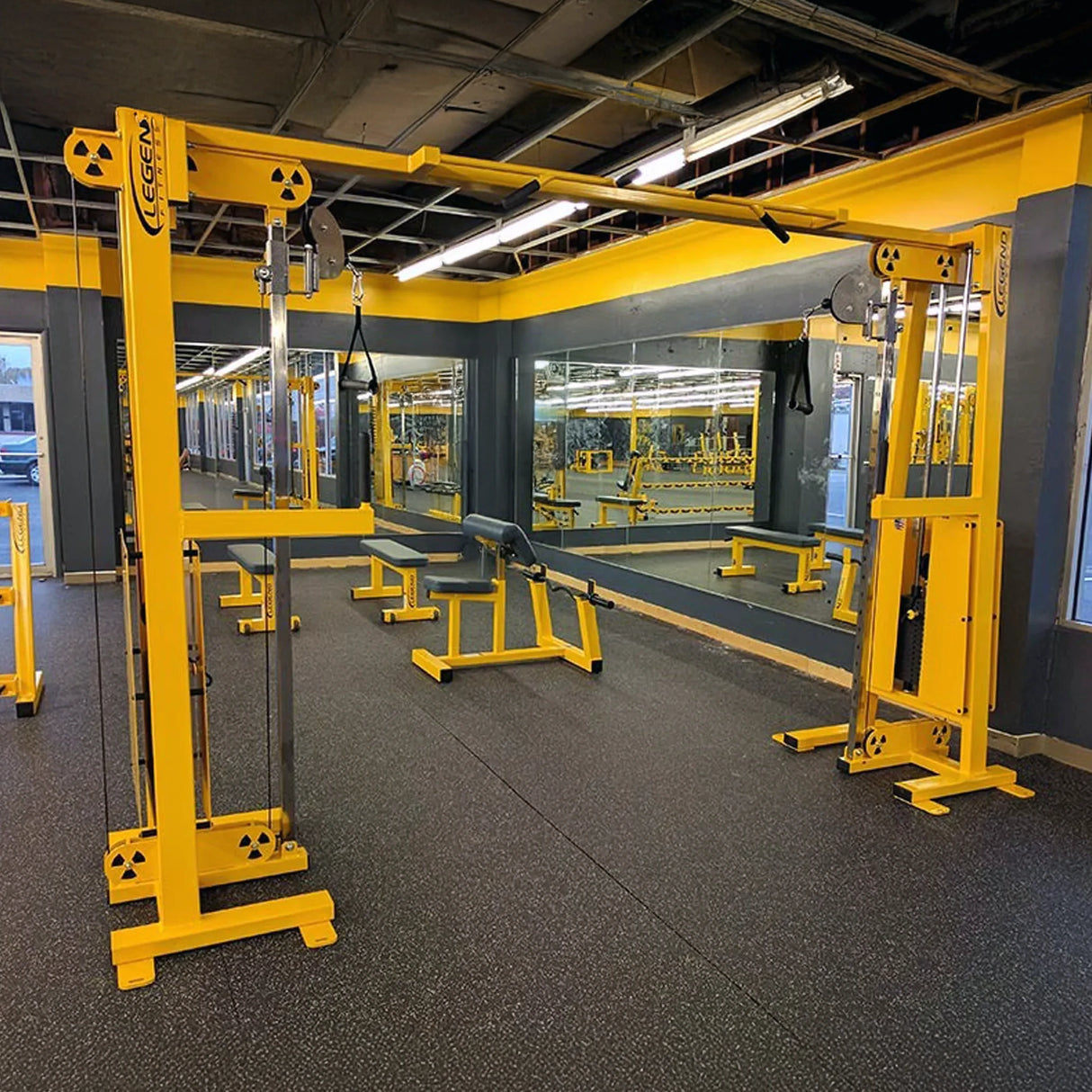 A brightly lit gym features Legend Fitness 954 Cable Crossover with Adjustable Pulleys, benches, and mirrored walls reflecting the yellow and black equipment. The floor is covered with rubberized black mats.