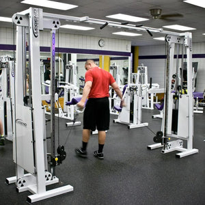 A man in a red shirt and black shorts uses the Legend Fitness 954 Cable Crossover with Adjustable Pulleys at a gym equipped with mirrors, a wall clock, and various exercise machines.