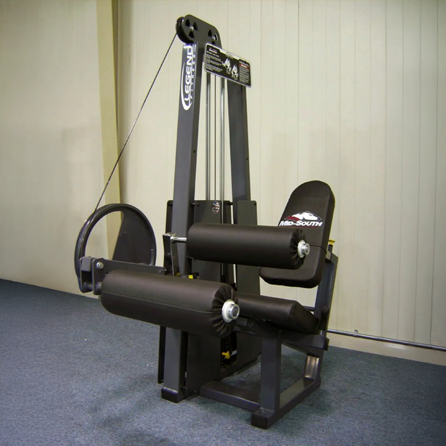 The Legend Fitness 956 Seated Leg Curl Machine by Legend Fitness features padded rollers and an adjustable seat. It is black and positioned on a blue carpeted floor against a beige paneled wall in the gym.