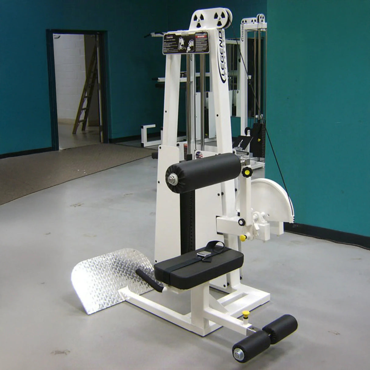 A Legend Fitness 957 Low Back / Ab Combo Machine with padded seats and rollers stands in a gym with teal walls, gray flooring, a metal footplate on the left, mirrors, and other equipment visible in the background.