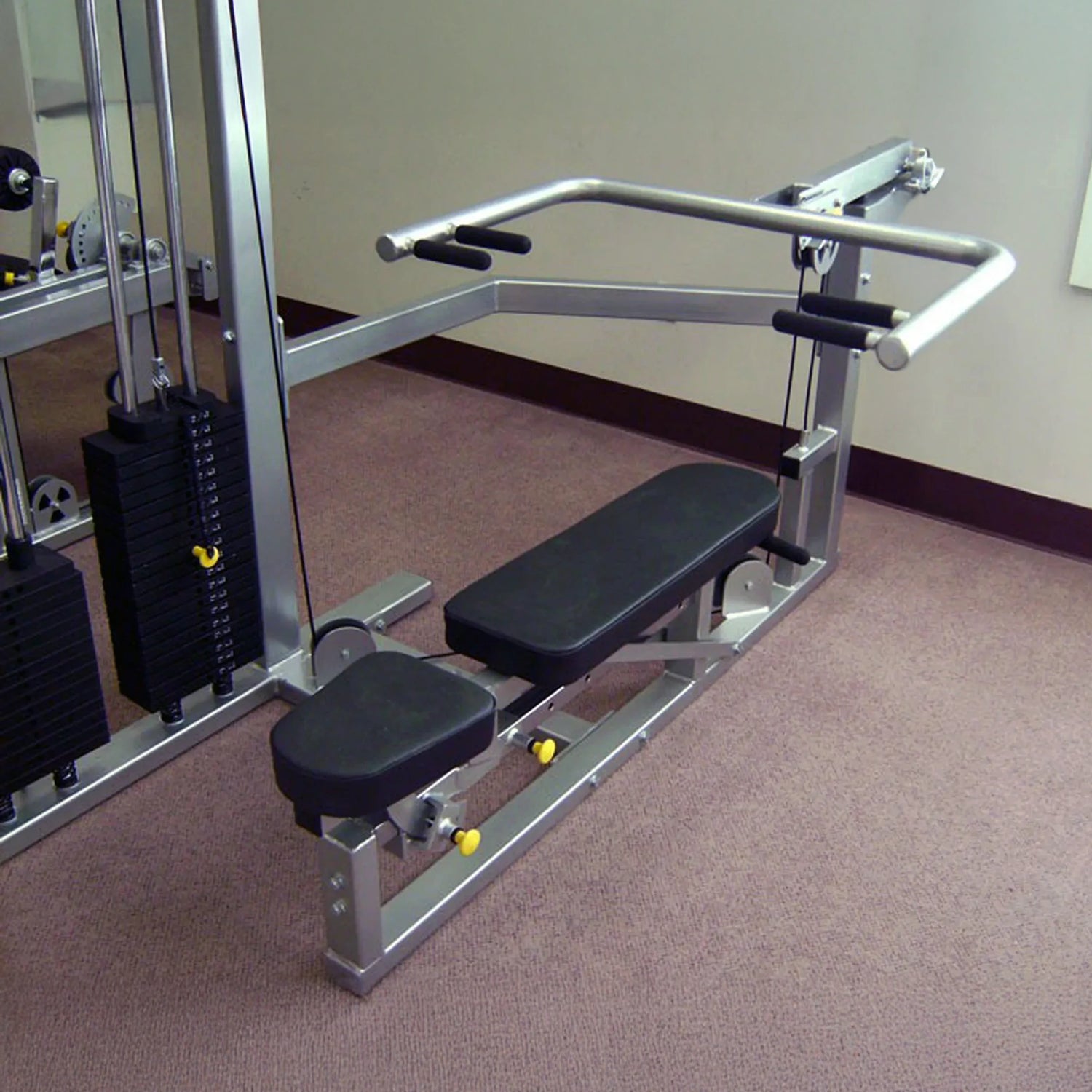 The Legend Fitness 958 Four-Stack Jungle Gym by Legend Fitness is a strength training machine with a padded bench and arm handles, designed for gyms and positioned on a carpeted floor next to a wall.