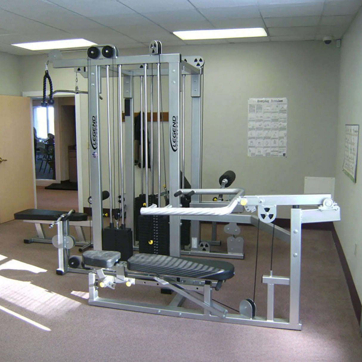 The Legend Fitness 958 Four-Stack Jungle Gym by Legend Fitness, featuring multiple attachments and cables, is set up in a carpeted room with light-colored walls, fitness charts, and an open door visible in the background.