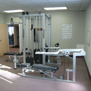 The Legend Fitness 958 Four-Stack Jungle Gym by Legend Fitness, featuring multiple attachments and cables, is set up in a carpeted room with light-colored walls, fitness charts, and an open door visible in the background.