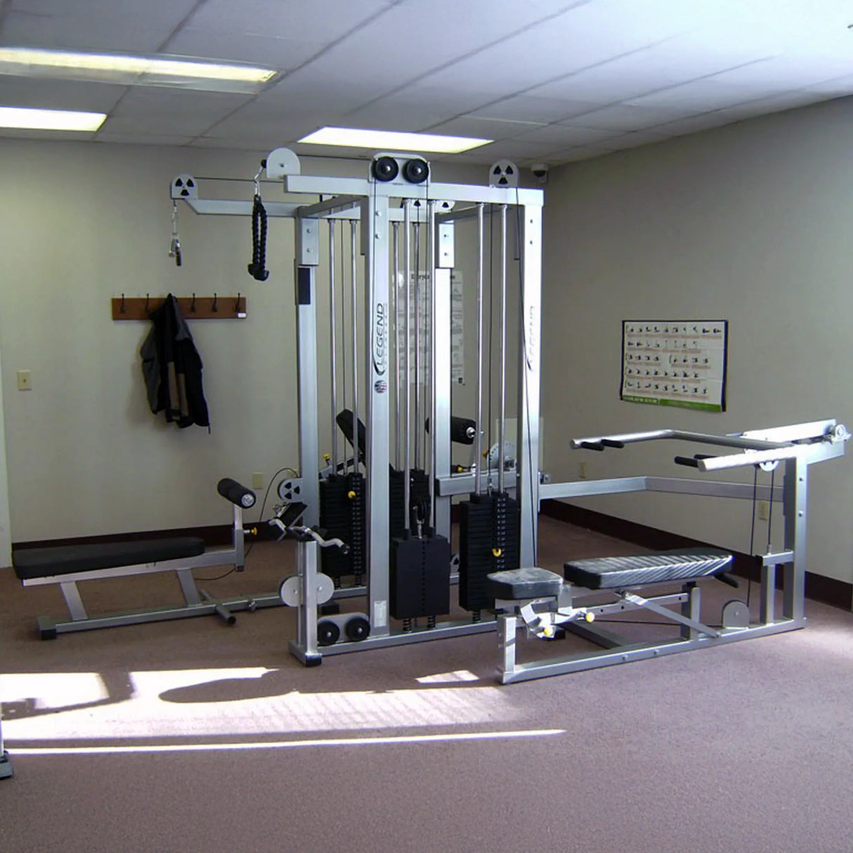 A small gym room features the Legend Fitness 958 Four-Stack Jungle Gym, padded benches, coat hooks with hanging clothes, beige carpet, and a workout diagram on the wall under fluorescent lights.