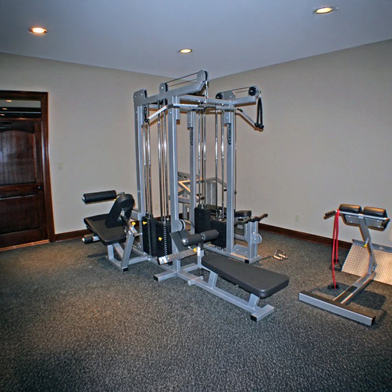 The room features a Legend Fitness 958 Four-Stack Jungle Gym with padded benches and attached cables, plus a hyperextension bench with red resistance bands on carpeted flooring, beige walls, and recessed lighting.