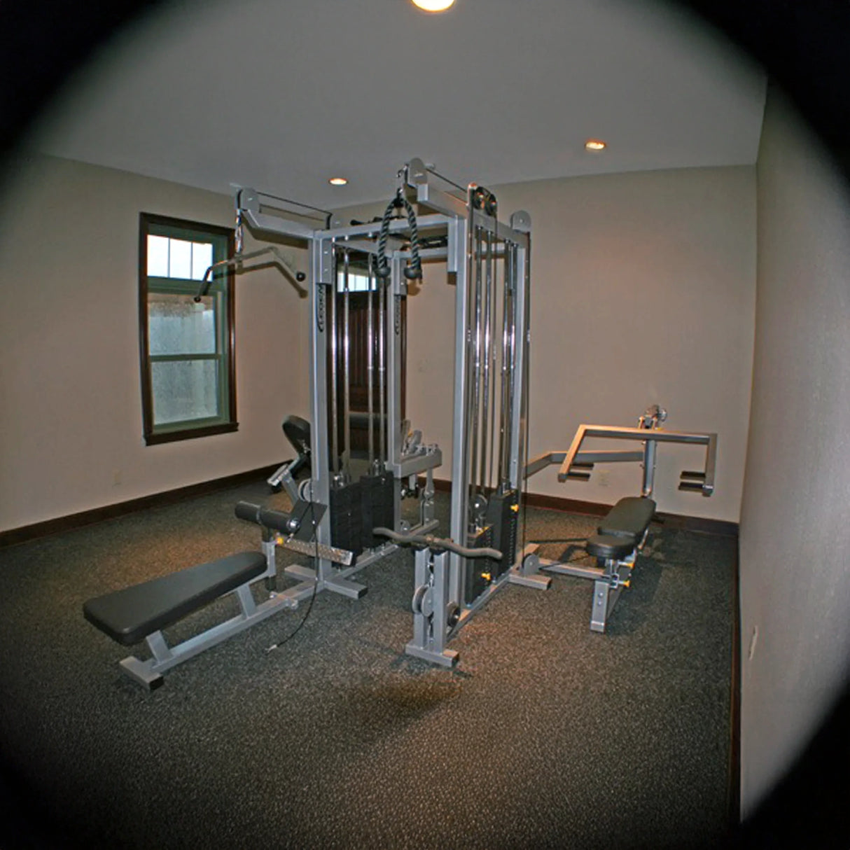 A small gym room with beige walls features the Legend Fitness 958 Four-Stack Jungle Gym and benches on a dark carpeted floor. Two windows and ceiling lights provide a bright, simple workout space.