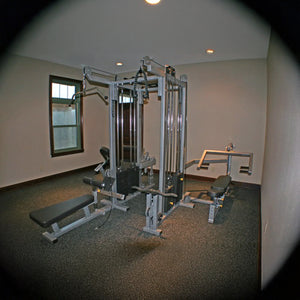 A small gym room with beige walls features the Legend Fitness 958 Four-Stack Jungle Gym and benches on a dark carpeted floor. Two windows and ceiling lights provide a bright, simple workout space.