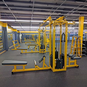 A spacious, well-lit gym featuring the Legend Fitness 959 Eight-Stack Jungle Gym in yellow and black, with benches, weight machines, and wall mirrors; the area is empty under bright overhead fluorescent lights.