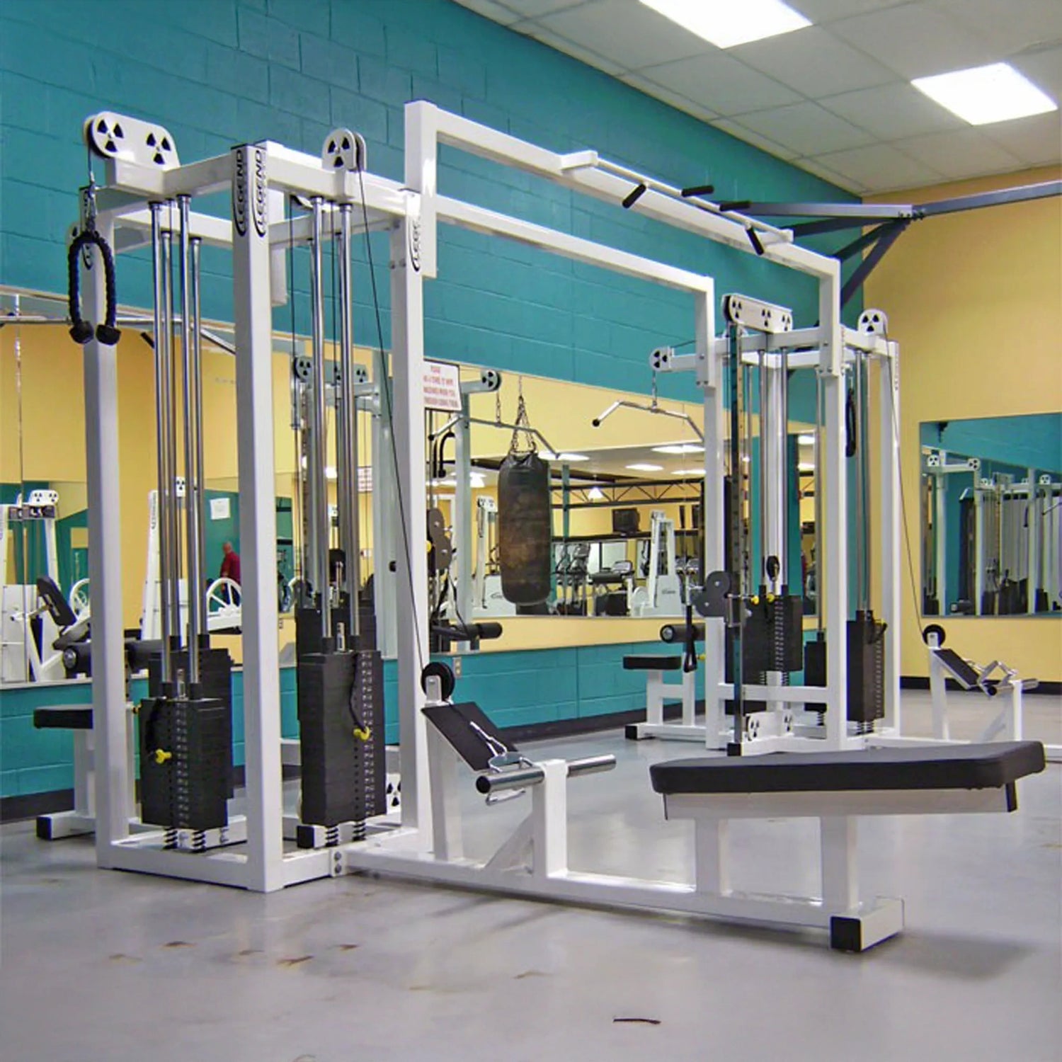 A gym featuring the Legend Fitness 959 Eight-Stack Jungle Gym by Legend Fitness, with weight stations like a cable crossover and seated row, bright overhead lighting, large mirrors, teal accent walls, and a punching bag in the background.