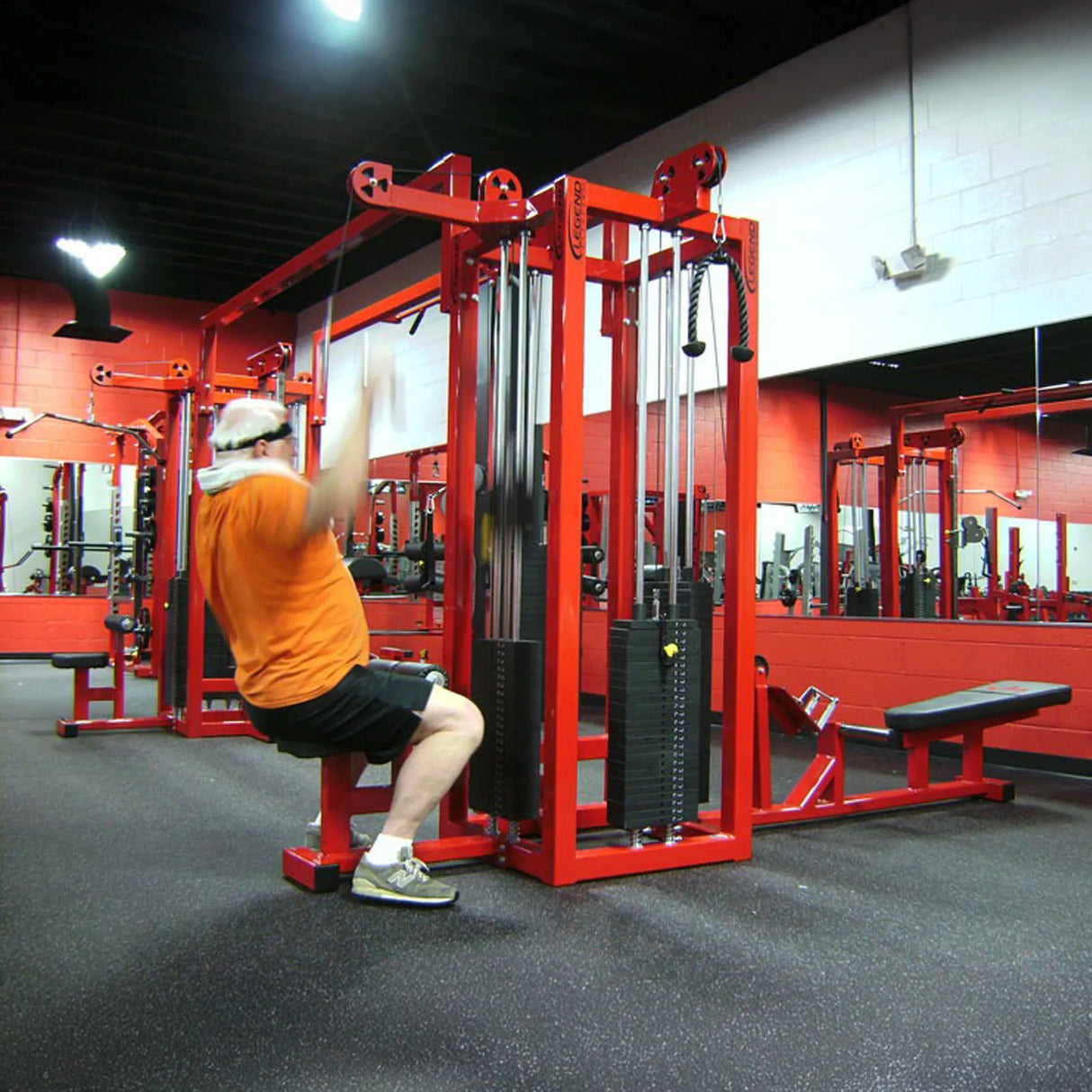 A person in an orange shirt performs a lat pulldown using a red Legend Fitness 959 Eight-Stack Jungle Gym in a facility with red and black equipment and a large wall mirror.