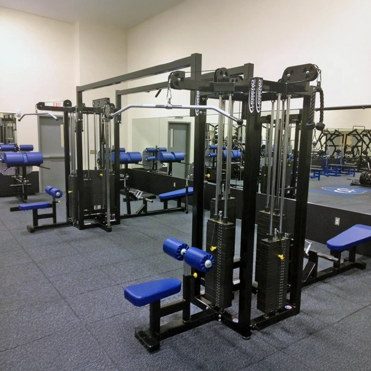 A clean gym features Legend Fitness 959 Eight-Stack Jungle Gym weight machines with blue padding on a carpeted floor. Large mirrors line the back wall, reflecting the Legend Fitness equipment and other machines.