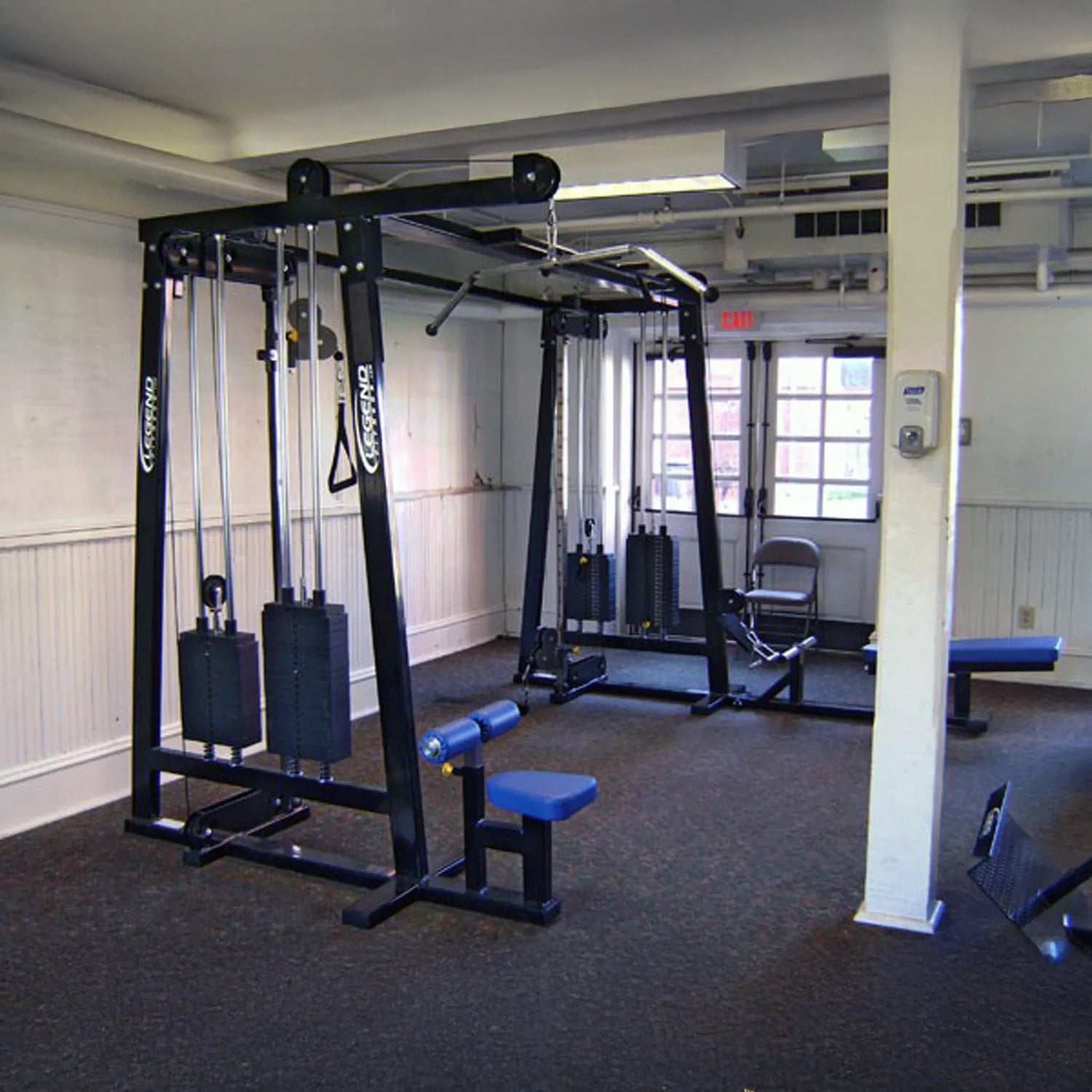 A compact gym room featuring the Legend Fitness 960 Cable Crossover Plus Machine, an adjustable bench, white walls, carpeted floor, and windows for natural light. The space has minimal equipment and a central pillar.