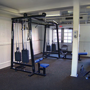 A compact gym room featuring the Legend Fitness 960 Cable Crossover Plus Machine, an adjustable bench, white walls, carpeted floor, and windows for natural light. The space has minimal equipment and a central pillar.