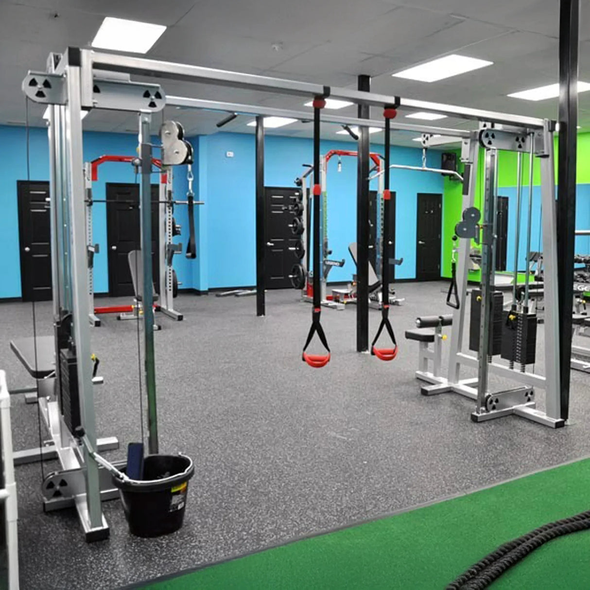 A modern gym featuring the Legend Fitness 960 Cable Crossover Plus Machine and other fitness equipment sits on a gray rubber floor, with bright blue and green walls and several black doors in the background.