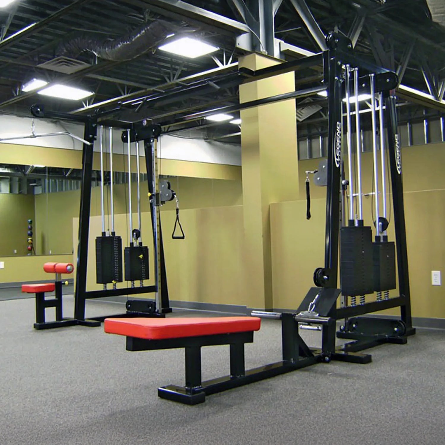 The Legend Fitness 960 Cable Crossover Plus Machine by Legend Fitness features red and black padded benches, set in an empty gym with beige walls, mirrors, and exposed ceiling pipes.