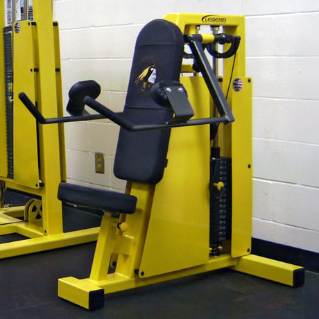 A Legend Fitness 962 Lateral Raise Machine with a padded seat, backrest, and arm pads is in a gym with black flooring and white walls. The yellow and black machine has weight plates visible on the side.