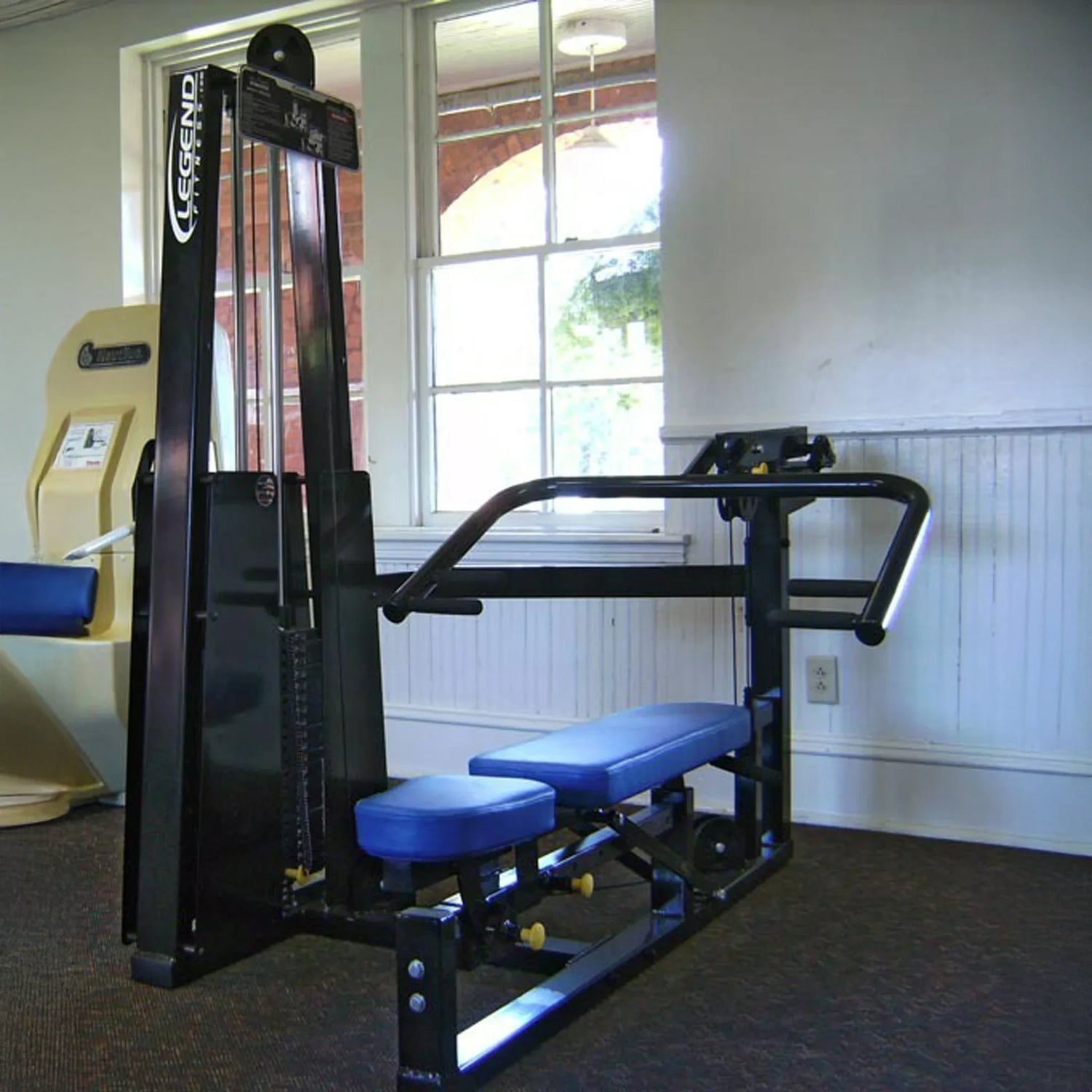 The Legend Fitness 963 Multi-Press Combo Machine, featuring blue padding and a black frame, is placed near a window in the fitness room. This Legend Fitness machine uses a weight stack and has handles and seat for upper body workouts.