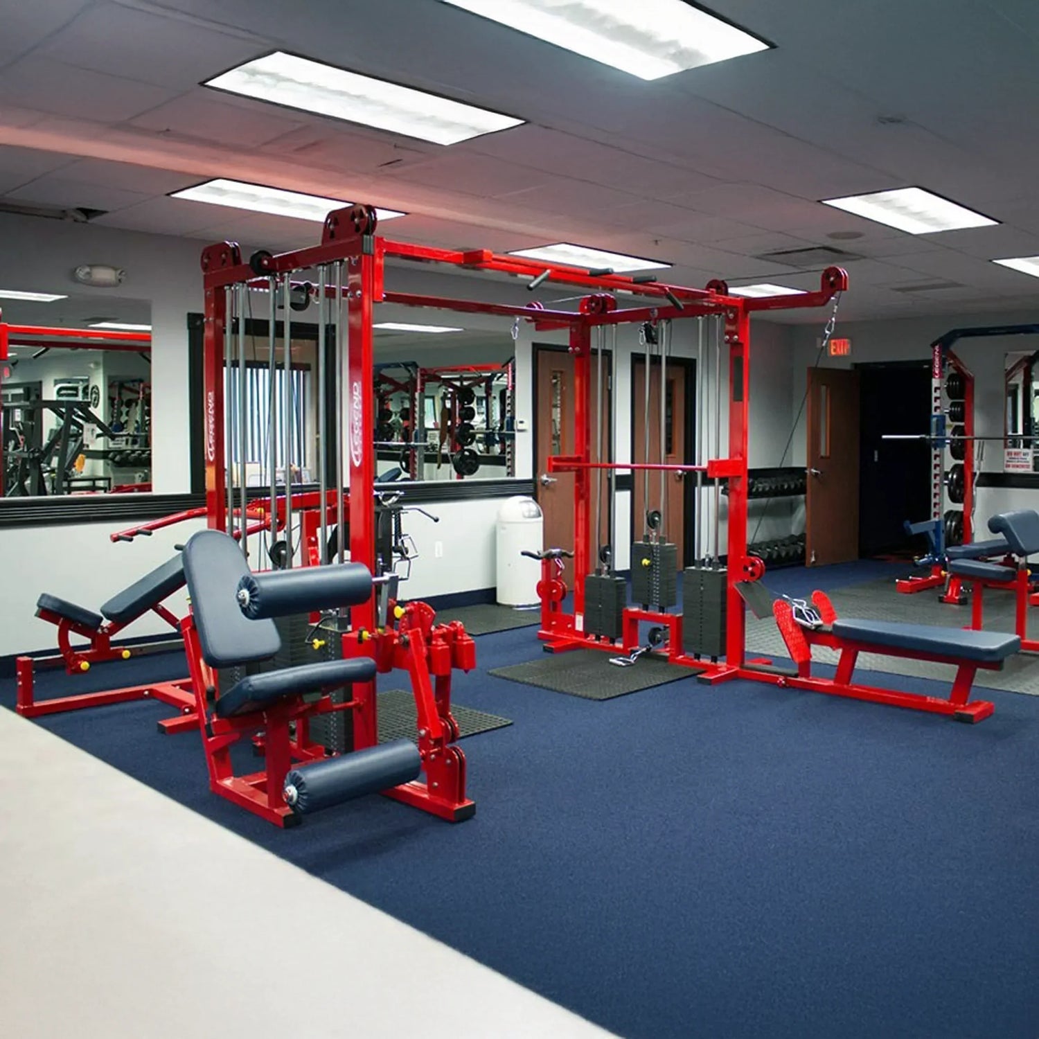 A gym with blue carpet features red Legend Fitness 965 Combo Jungle Machines, padded benches, wall mirrors, and fluorescent ceiling lights. A door and additional exercise equipment are visible in the background.