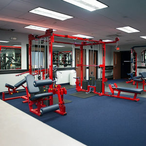 A gym with blue carpet features red Legend Fitness 965 Combo Jungle Machines, padded benches, wall mirrors, and fluorescent ceiling lights. A door and additional exercise equipment are visible in the background.