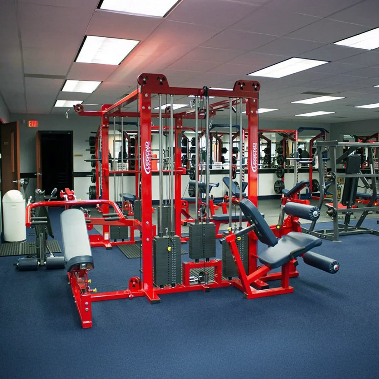 A modern gym with blue carpet flooring features the Legend Fitness 965 Combo Jungle Machine by Legend Fitness, with weight stacks, benches, and mirrors lining the walls under bright ceiling lights.