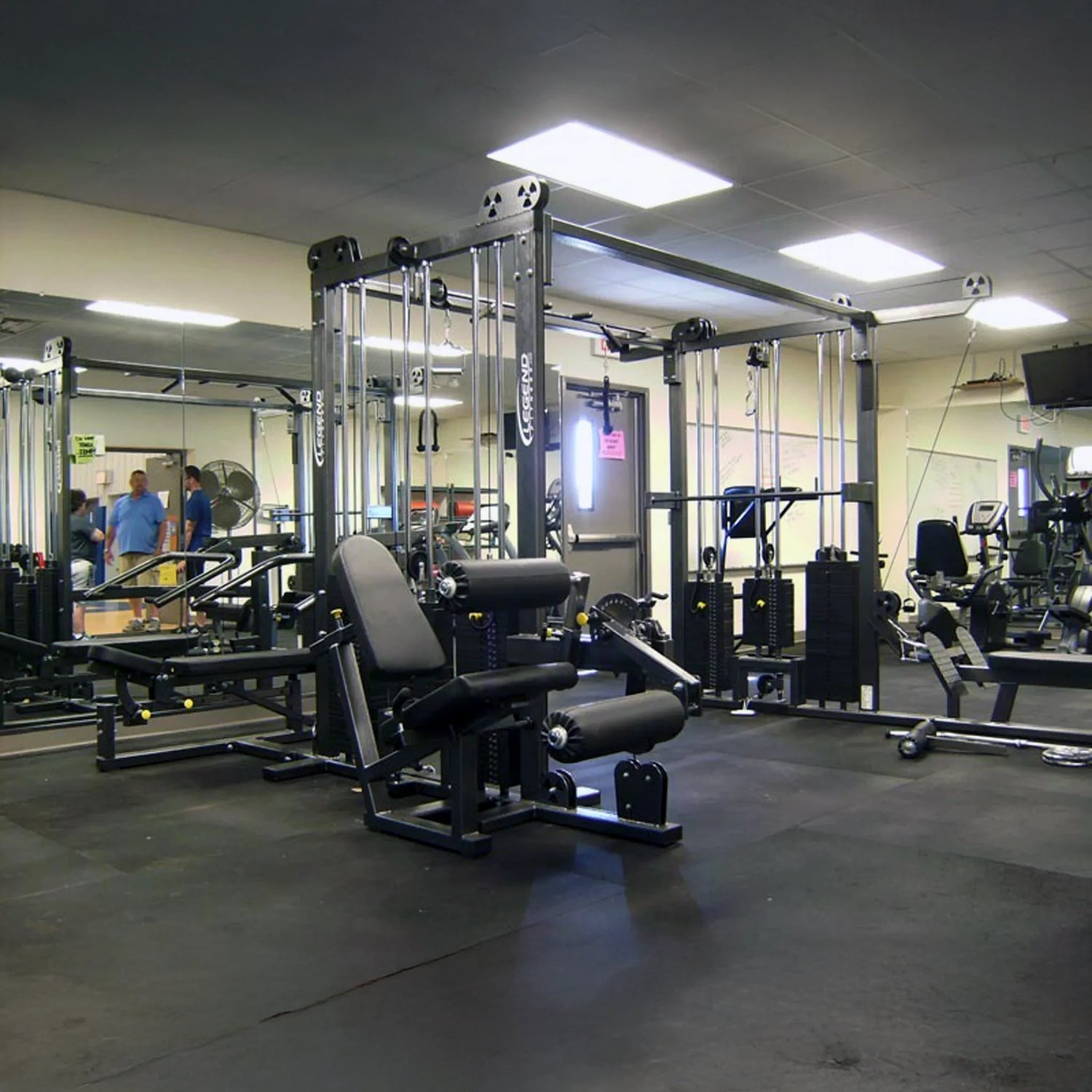 A gym interior features the Legend Fitness 965 Combo Jungle Machine among other exercise equipment. Large wall mirrors and black rubber flooring are visible, with two people near the mirrors in the background.