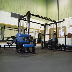 A gym featuring Legend Fitness 965 Combo Jungle Machines in blue and black on a rubber floor, with mirrors and bulletin boards on light brick walls, all brightly lit by fluorescent lights.