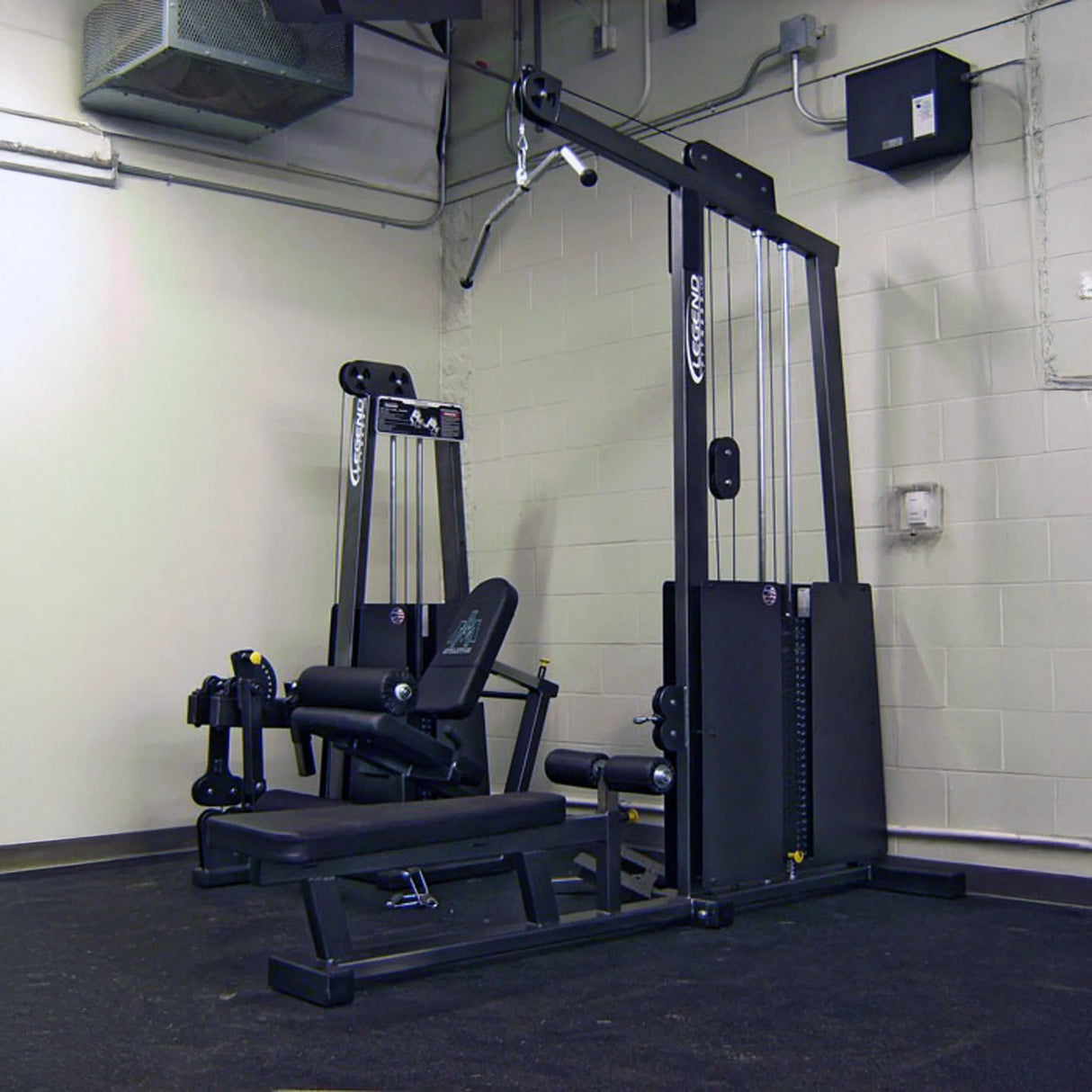 A gym corner with a Legend Fitness 971 Pro Seated Lat Pulldown / Low Row Machine by Legend Fitness sits against a light-colored wall on a rubber floor, with vents, electrical boxes, and exposed pipes on the ceiling and walls.