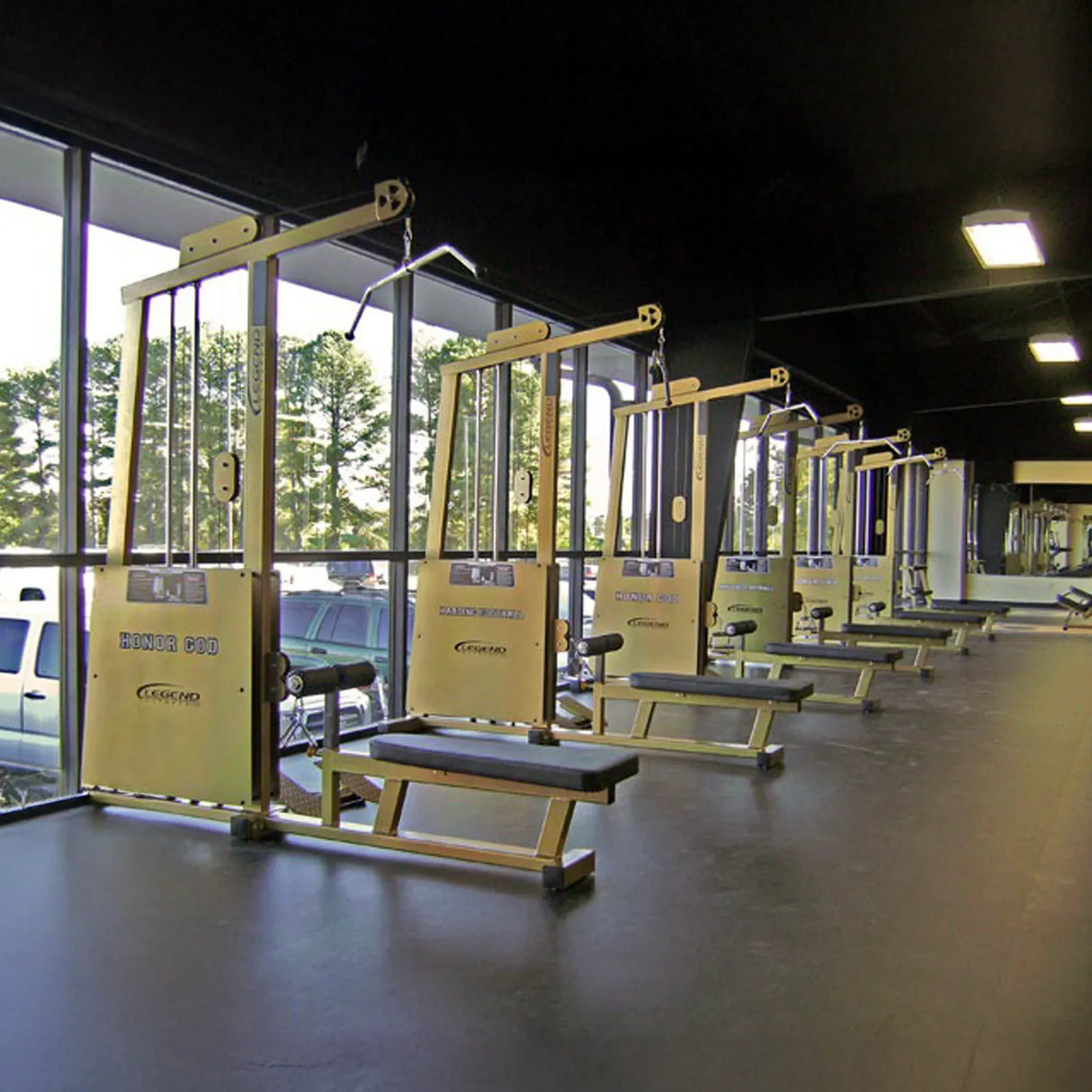 A row of Legend Fitness 971 Pro Seated Lat Pulldown / Low Row Machines is arranged by large windows in a bright, clean gym, with trees and parked cars visible outside. The space feels modern and spacious.