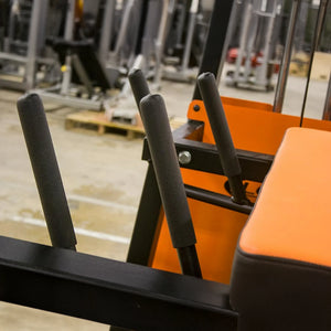 Close-up of foam-padded handles on the orange and black Legend Fitness 974 Pro Reverse Back Extension Machine, with other gym equipment blurred in the background.