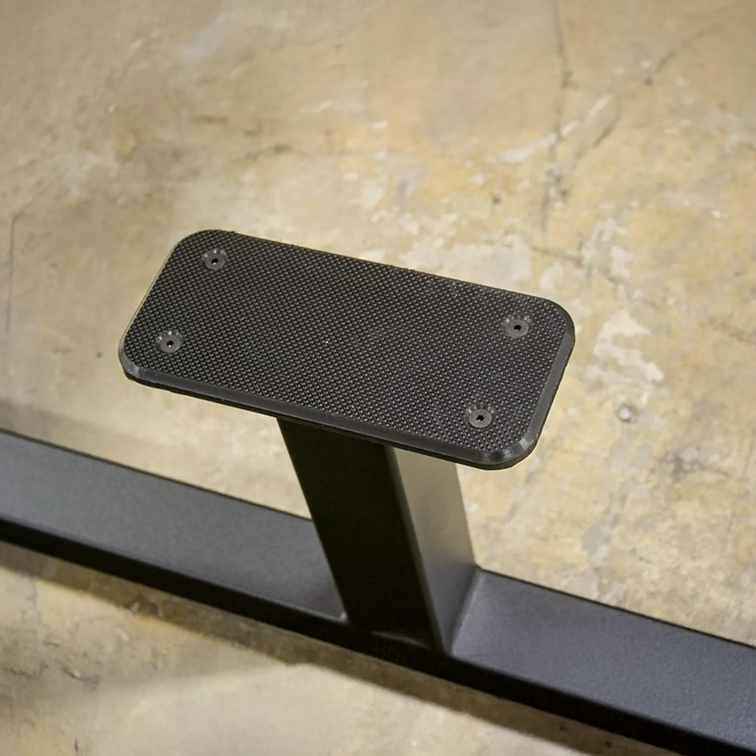 A close-up of the textured black metal footrest on the Legend Fitness 974 Pro Reverse Back Extension Machine, attached with four visible screws to a metal support on a concrete floor.
