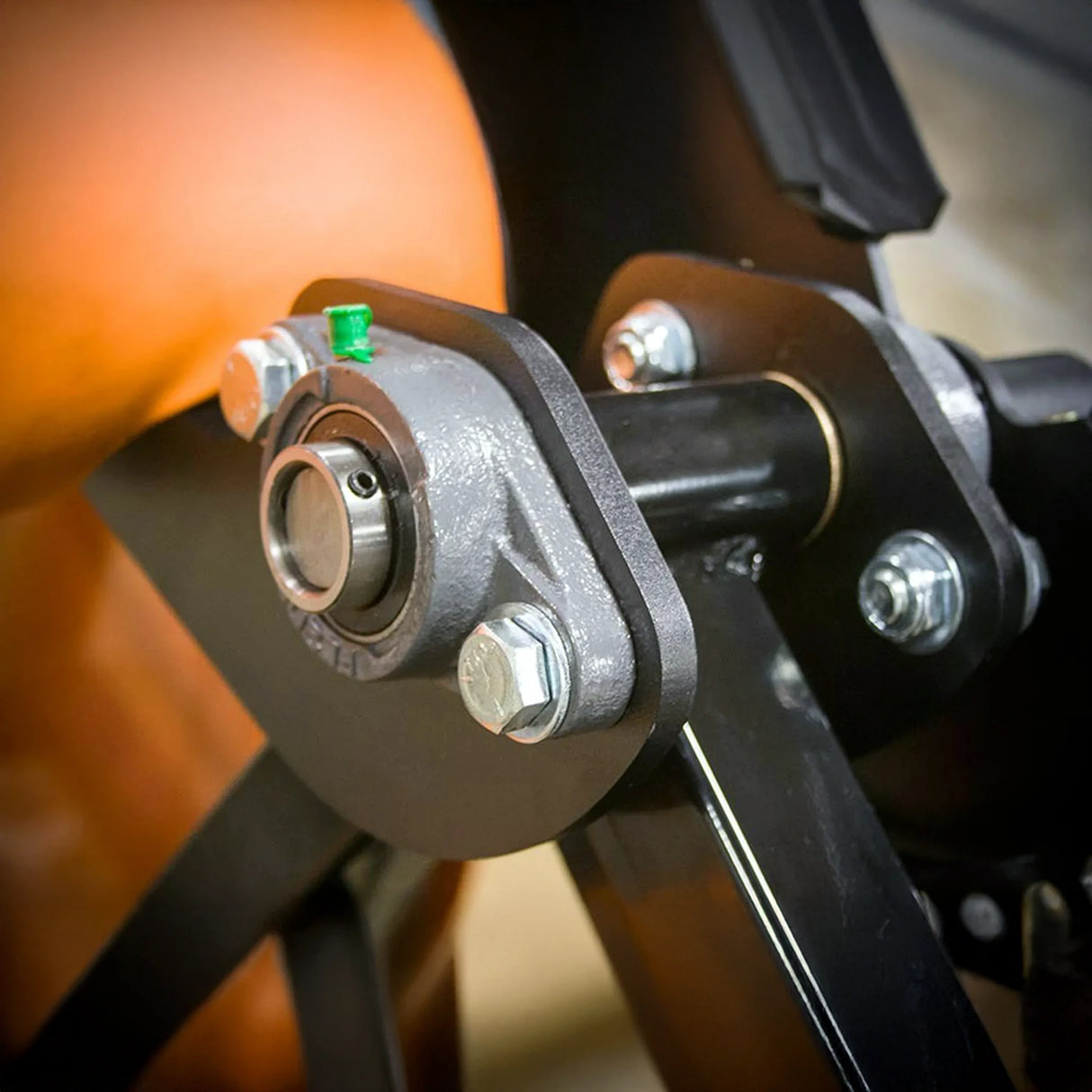Close-up of a metal mechanical joint with bolts, bearing, and green grease fitting attached to the black frame of the Legend Fitness 974 Pro Reverse Back Extension Machine by Legend Fitness. Part of an orange object appears in the background.