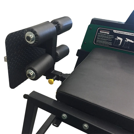 Close-up of the Legend Fitness 976-GH Reverse Back Extension/GHD Combo Machine, featuring black padded rollers, a diamond-plate footrest, part of a cushioned seat, visible adjustment knobs, and instruction labels on the Legend Fitness frame.