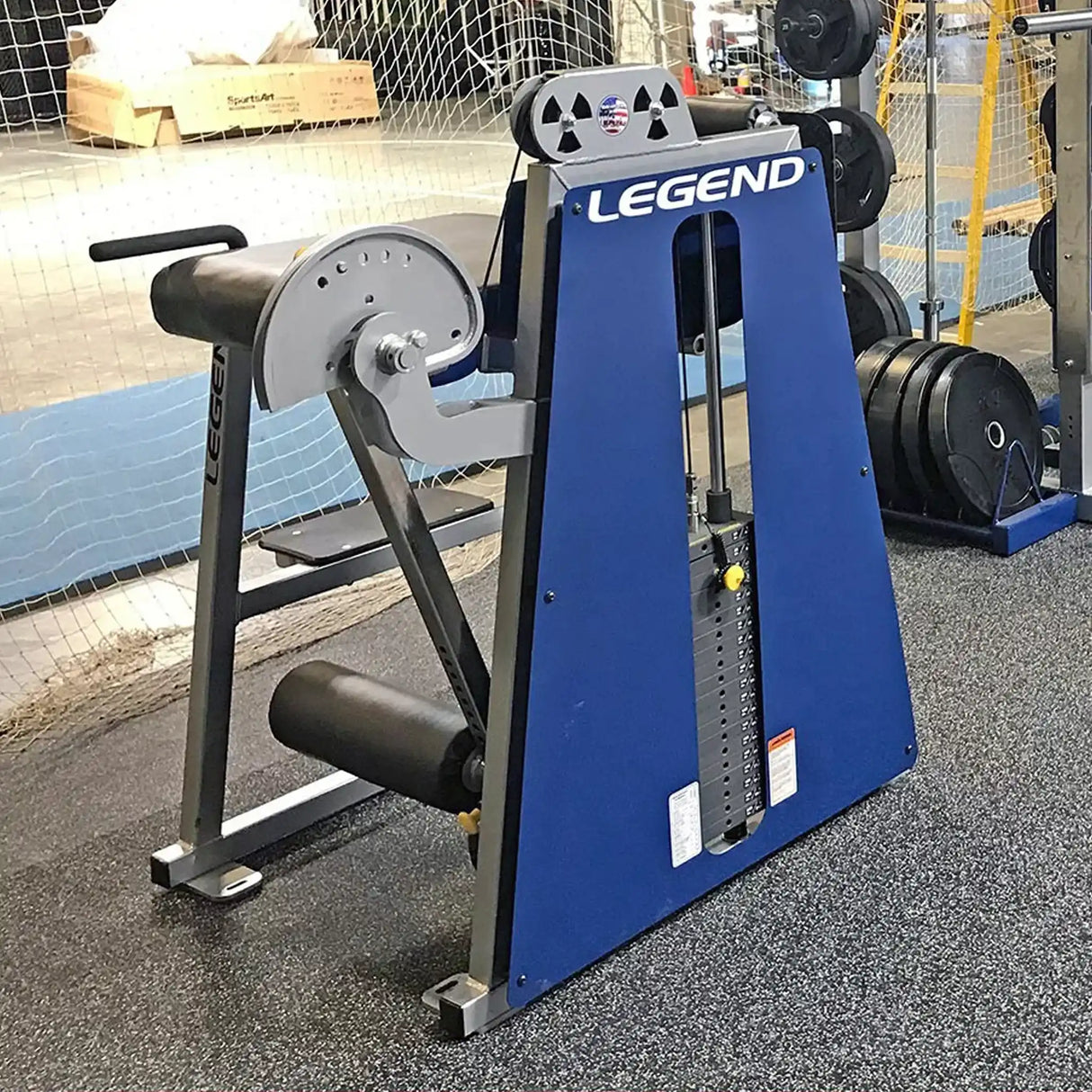 A Legend Fitness 976 Performance Reverse Back Extension Machine in blue is in a gym, featuring a padded seat, leg rollers, and an adjustable weight stack with a yellow pin; other gym equipment can be seen in the background.