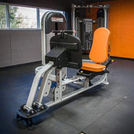 A white gym leg press machine with orange seat in a gym setting