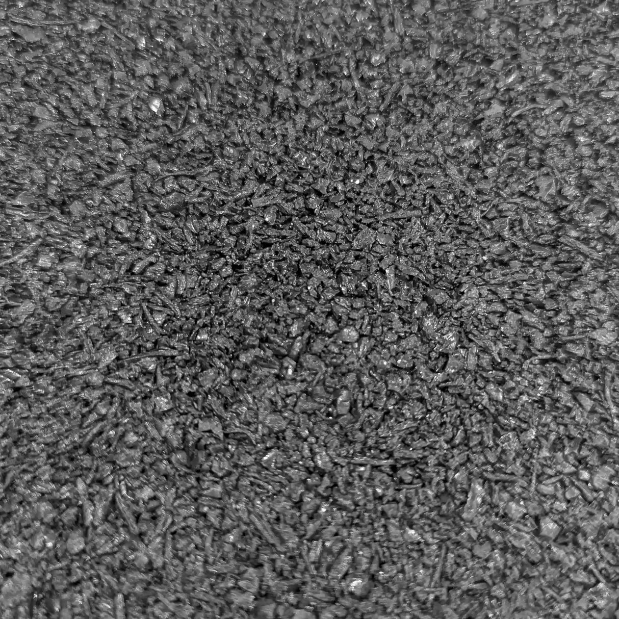 A close-up of ECORE Athletic's ECORE Rubber Pavers (1-3/4", 44.5 mm) shows a rough, textured dark gray surface with scattered small pebbles and irregular patterns, resembling asphalt or coarse concrete.