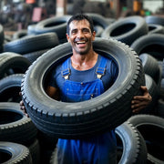 Smiling man holding a tire around his neck amidst stacked tires in a warehouse setting