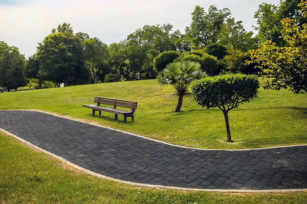 A black rubber paver footpath running through a green park with trees and a bench