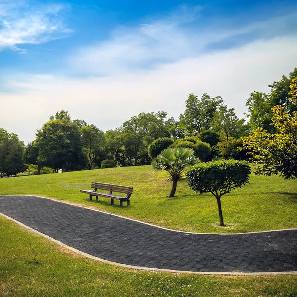 A black rubber paver footpath running through a green park with trees and a bench