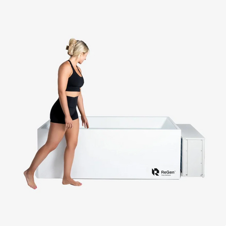 A rectangular cold plunge with a white exterior and a white external chiller unit on the right side of the plunge. There is a blonde woman walking down the side of the plunge looking as if she's about to get into it.