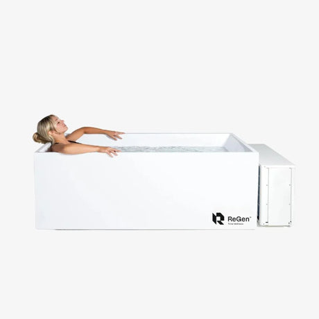 A rectangular cold plunge filled with water with a white exterior and a white external chiller unit on the right side of the plunge. In the plunge is a blonde woman lying down with her eyes closed.