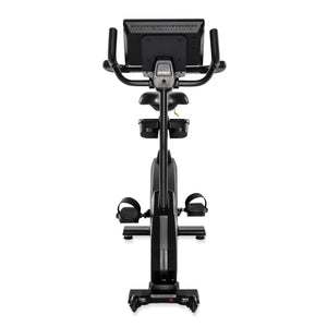 Rear view of the black Spirit Fitness CU1000ENT Upright Bike showing the back of the console, seat with dual accessory/water bottle holders below, pedals with adjustable straps, and the rear base with built-in transport wheels.